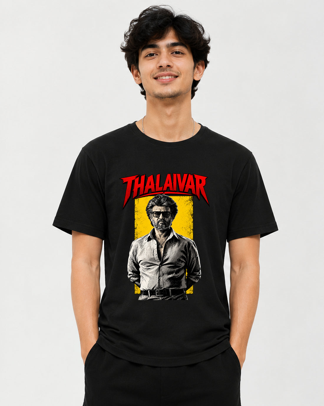 A man wearing a black graphic t-shirt featuring a red 'THALAIVAR' logo and a stylized portrait of Thalaivars on a yellow background, set against a plain light gray studio backdrop.