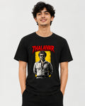 A man wearing a black graphic t-shirt featuring a red 'THALAIVAR' logo and a stylized portrait of Thalaivars on a yellow background, set against a plain light gray studio backdrop.