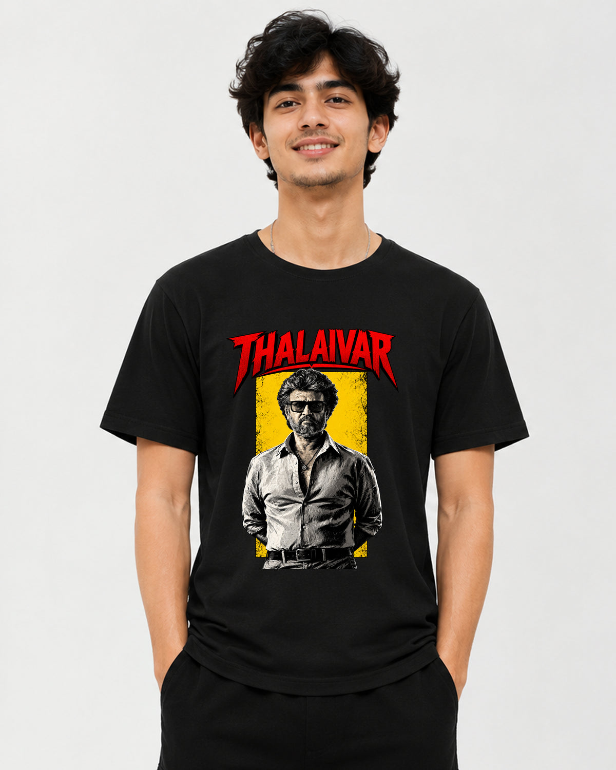 A man wearing a black graphic t-shirt featuring a red 'THALAIVAR' logo and a stylized portrait of Thalaivars on a yellow background, set against a plain light gray studio backdrop.